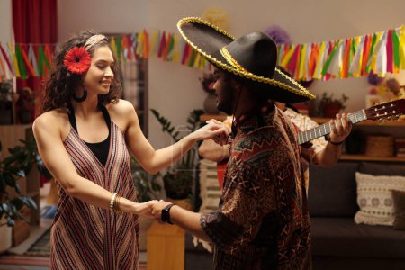 Photo for Happy young woman in ethnic dress holding her boyfriend in sombrero by hands during dance in home environment while enjoying party - Royalty Free Image