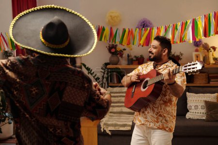 Téléchargez les photos : Jeune homme heureux avec guitare espagnole debout devant un couple dansant et jouant de l'instrument de musique à cordes tout en appréciant la fête à la maison - en image libre de droit