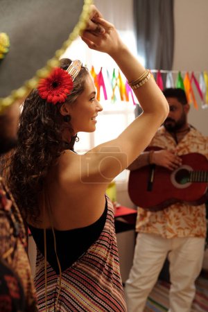 Foto de Feliz joven morena con flor roja en el pelo cogida de la mano de su novio durante la danza dinámica contra el músico tocando la guitarra - Imagen libre de derechos