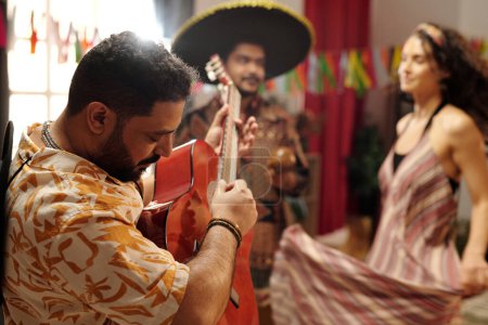 Téléchargez les photos : Jeune musicien barbu en tenue nationale jouant de la guitare devant la caméra pendant que l'homme et la femme dynamiques dansent à la fête à la maison - en image libre de droit