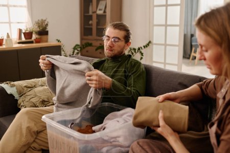 Téléchargez les photos : Jeune homme tenant le vieux pull gris et le regardant assis sur le canapé devant sa femme et sélectionnant des vêtements pour le don - en image libre de droit