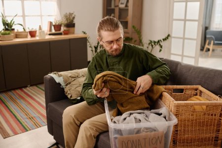 Photo for Bearded blond man in dark green shirt and beige pants selecting old clothes for donation center while sitting on couch in living room - Royalty Free Image