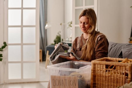 Téléchargez les photos : Femme blonde volontaire pliant et emballant de vieux vêtements dans une boîte en plastique et un panier à la maison tout en préparant des vêtements pour le centre de dons - en image libre de droit