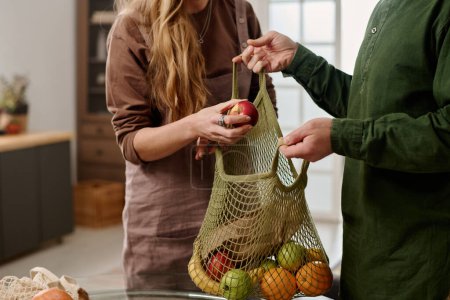 Photo for Cropped shot of young woman putting red apple into shopping bag with various fruits held by her husband while both standing in front of camera - Royalty Free Image