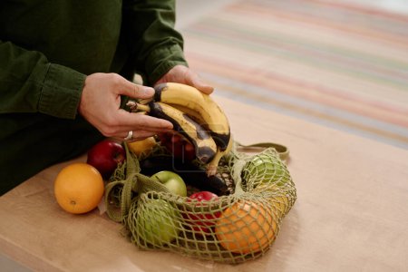 Foto de Manos de hombre joven irreconocible sosteniendo racimo de plátanos podridos sobre la bolsa de compras con manzanas maduras frescas y naranjas en la mesa - Imagen libre de derechos