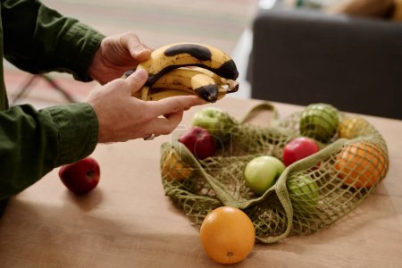 Téléchargez les photos : Les mains d'un homme méconnaissable sélectionnant des fruits dans un sac à provisions tout en restant près du comptoir de la cuisine et en tenant des bananes pourries - en image libre de droit