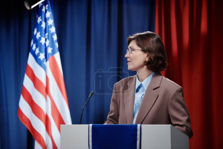 Téléchargez les photos : Femme politique brune mature regardant de côté tout en restant près de la plate-forme, parlant au micro et faisant une présentation - en image libre de droit