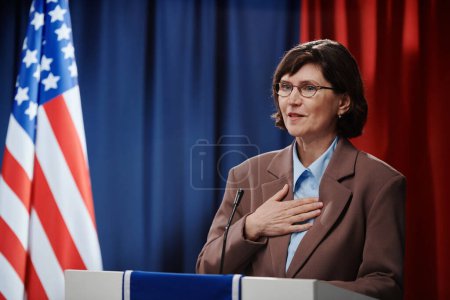 Téléchargez les photos : Femme mûre candidate au poste de présidente en gardant la main sur la poitrine et en regardant l'électorat tout en parlant au microphone - en image libre de droit