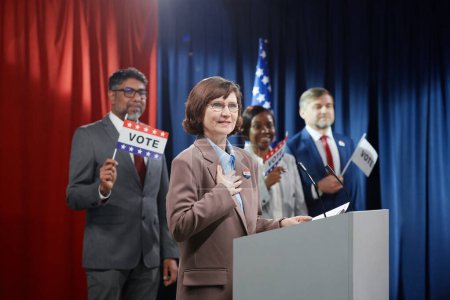 Téléchargez les photos : Femme souriante mature gardant la main sur la poitrine et regardant l'électorat tout en se tenant près de la plate-forme et parlant au microphone - en image libre de droit
