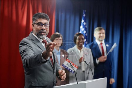 Téléchargez les photos : Un homme politique multiethnique confiant en tenue de cérémonie prononce un discours pendant la campagne électorale tout en se tenant debout contre ses collègues - en image libre de droit