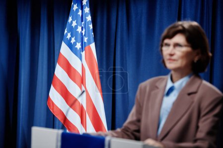 Foto de Bandera estadounidense en plataforma con micrófono y mujer política madura desenfocada en ropa formal de pie frente al público durante el discurso - Imagen libre de derechos