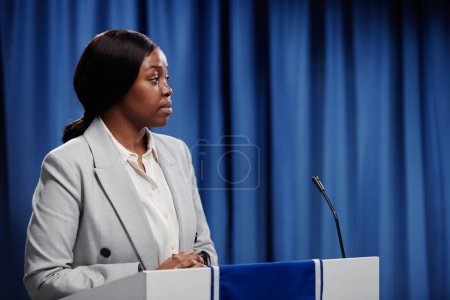 Foto de Joven mujer política mirando a su oponente durante el debate político o en la conferencia mientras está de pie en la plataforma con micrófono - Imagen libre de derechos