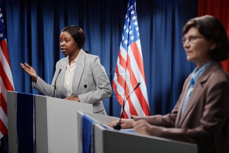 Foto de Una joven delegada confiada en pie de plataforma contra la bandera estadounidense en la sala de conferencias y respondiendo a la pregunta de la prensa - Imagen libre de derechos