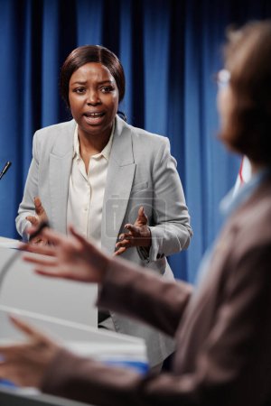 Téléchargez les photos : Jeune femme afro-américaine agacée déléguée debout par tribune et regardant son adversaire pendant le débat et criant à elle - en image libre de droit