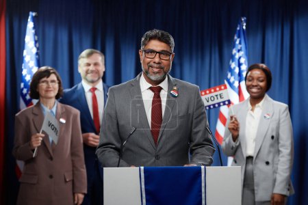 Téléchargez les photos : Candidat masculin multiethnique mature faisant un discours pendant la campagne électorale et regardant la caméra tout en se tenant devant le public - en image libre de droit