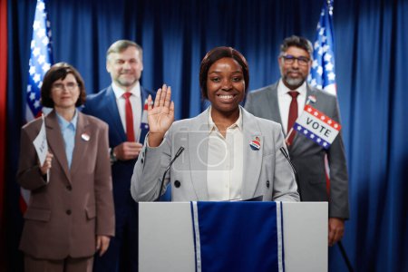 Foto de Joven y alegre mujer política saludando con la mano o mostrando gesto de voto y mirando a la cámara mientras está de pie en la plataforma contra los delegados - Imagen libre de derechos