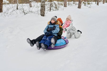 Foto de Dos niños afectuosos felices deslizándose colina abajo en tubo de nieve mientras linda chica en ropa de invierno empujándolos durante el juego en el bosque de invierno - Imagen libre de derechos