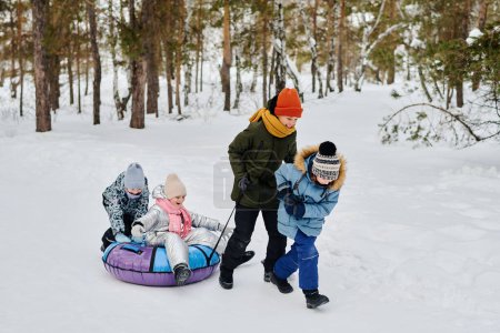 Téléchargez les photos : Deux garçons heureux en bonneterie et vestes d'hiver tirant tube à neige avec l'une des deux filles tout en descendant la dérive des neiges dans le parc ou la forêt - en image libre de droit