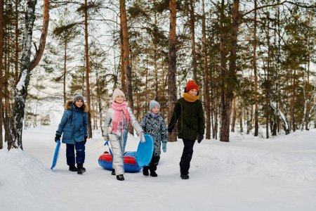 Téléchargez les photos : Quatre enfants interculturels joyeux en vestes d'hiver chaudes et bonnets tirant des tubes à neige et portant des diapositives tout en marchant le long de la route - en image libre de droit