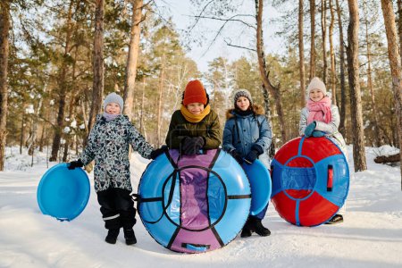 Foto de Varios niños interculturales felices en ropa de invierno sosteniendo tubos de nieve y diapositivas y mirando a la cámara mientras juega al aire libre en el día de invierno - Imagen libre de derechos