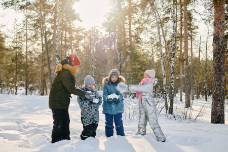 Téléchargez les photos : Enfants mignons en vêtements d'hiver mettre de la neige sur les mains de leurs amis tout en se tenant dans la dérive des neiges dans le parc et faire bonhomme de neige ensemble - en image libre de droit
