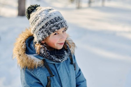 Téléchargez les photos : Mignon garçon souriant en bonnet gris tricoté et veste d'hiver bleue regardant la caméra tout en marchant le long de la route dans le parc ou la forêt - en image libre de droit