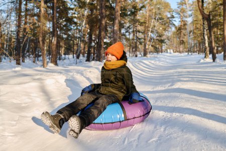 Téléchargez les photos : Joyeux garçon mignon en veste d'hiver chaude, bonnet et écharpe assis sur un tube à neige tout en descendant la colline dans un environnement naturel - en image libre de droit