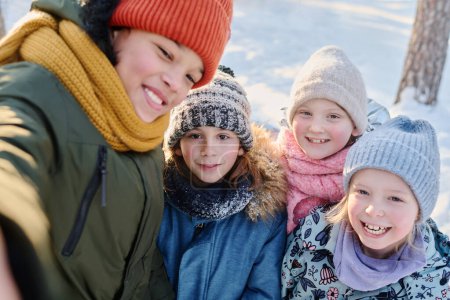 Foto de Uno de los cuatro niños felices tomando selfie en el teléfono inteligente mientras está de pie junto a sus amigos alegres que usan ropa de invierno caliente - Imagen libre de derechos