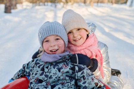Téléchargez les photos : Deux jolies filles avec des sourires de dents regardant la caméra tandis que l'un d'eux assis sur le tube à neige devant son adorable ami - en image libre de droit