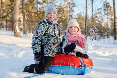 Téléchargez les photos : Deux filles riantes avec un tube à neige debout sur les genoux sur la route et regardant la caméra tout en profitant de la promenade le week-end d'hiver dans le parc - en image libre de droit