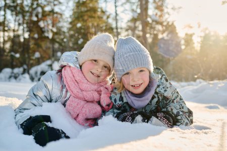Téléchargez les photos : Jolies petites filles en vêtements d'hiver regardant la caméra avec des sourires lors d'une journée d'hiver ensoleillée dans un environnement naturel tout en s'amusant dans la dérive des neiges - en image libre de droit