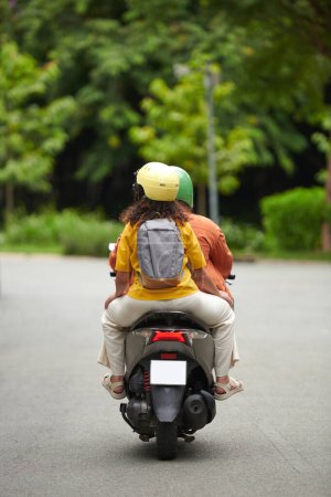 Foto de Vista trasera de una mujer joven con mochila sentada en motocicleta y sujetada por su esposo o novio conduciendo bicicleta - Imagen libre de derechos