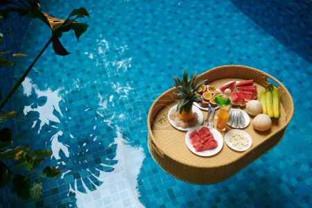 Téléchargez les photos : Grand angle de plateau avec divers fruits frais et jus debout sur la surface de l'eau bleue transparente de la piscine par hôtel de luxe - en image libre de droit