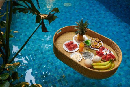 Téléchargez les photos : Grand angle d'eau bleue dans la piscine par hôtel luxueux et plateau avec assortiment de fruits exotiques frais et verre de jus d'orange - en image libre de droit