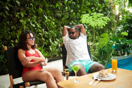 Téléchargez les photos : Young restful intercultural couple in sunglasses sitting by poolside at summer resort, sunbathing and enjoying sunny day - en image libre de droit
