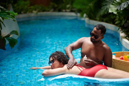 Téléchargez les photos : Young woman relaxing on air mattress on surface of blue water in swimming pool while her boyfriend applying lotion on her back - en image libre de droit