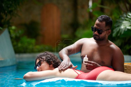 Foto de Idyllic scene of young serene couple spending time in swimming pool with blue water while guy applying sunblock lotion on back of girl - Imagen libre de derechos