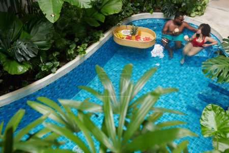 Téléchargez les photos : High angle of young restful couple sitting in blue water of swimming pool by luxurious hotel and having tropical drinks from calabashes - en image libre de droit