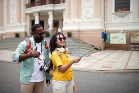 Téléchargez les photos : Young woman holding selfie stick with smartphone while taking picture of herself and her husband while both standing by historical building - en image libre de droit