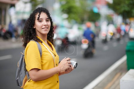 Téléchargez les photos : Young smiling brunette female traveler with photo camera in hands looking at you while standing by road in the street - en image libre de droit