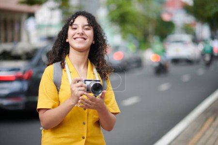 Téléchargez les photos : Young cheerful woman in casualwear looking at camera while standing in the street and taking shot of city sights during excursion - en image libre de droit