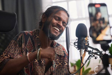 Téléchargez les photos : Happy young Indian man keeping eyes closed while speaking or singing in microphone during livestream for online audience in studio - en image libre de droit