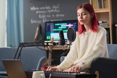 Téléchargez les photos : Pretty teenage girl with red hair playing electronic synthesizer by desk and singing in microphone while performing new song and recording it - en image libre de droit