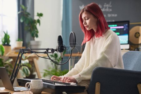 Téléchargez les photos : Pretty teenage girl in casualwear playing electric piano in home studio while standing in front of laptop and recording new music - en image libre de droit
