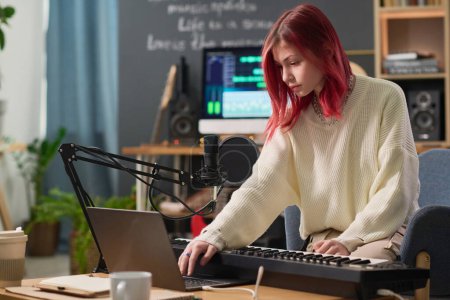 Téléchargez les photos : Cute teenage girl bending over desk with electric piano and laptop and looking at device screen while pressing key during sound recording - en image libre de droit