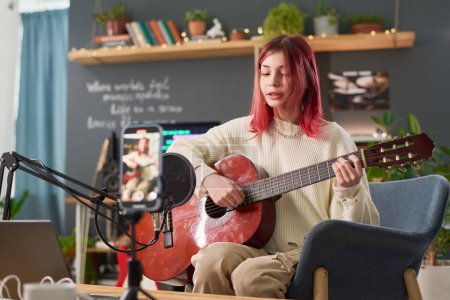Téléchargez les photos : Cute teenage girl in casualwear sitting in armchair in front of microphone, performing new song and playing acoustic guitar in home studio - en image libre de droit