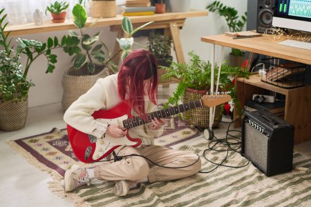 Téléchargez les photos : Teenage girl with red hair playing bass guitar while sitting on the floor in home studio, creating new music and recording it on computer - en image libre de droit