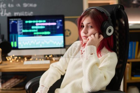 Photo for Cute serene teenage girl in headphones and white sweater listening to music in armchair against computer screen with audio software - Royalty Free Image