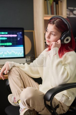 Téléchargez les photos : Serene teenager in casual attire and headphones sitting in armchair in front of camera in home studio and listening to her music - en image libre de droit