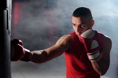 Young muscular man in red vest and boxing gloves hitting punching bag during training in gym or sports center with smoke on background